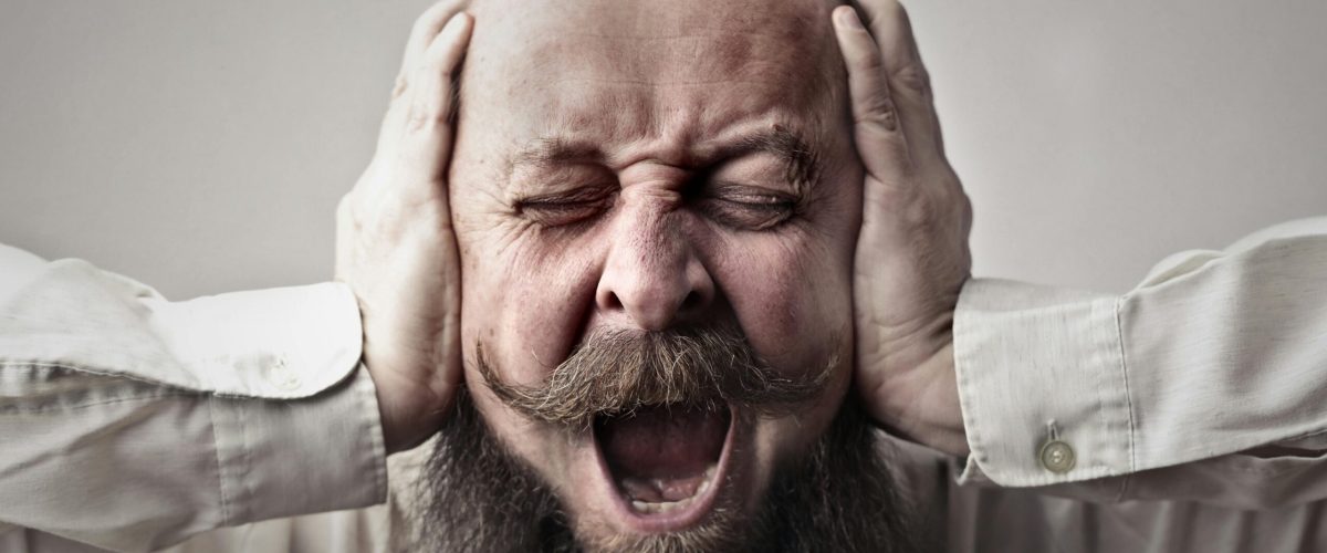 Portrait of a man with facial hair, covering his ears and screaming, expressing stress or frustration.